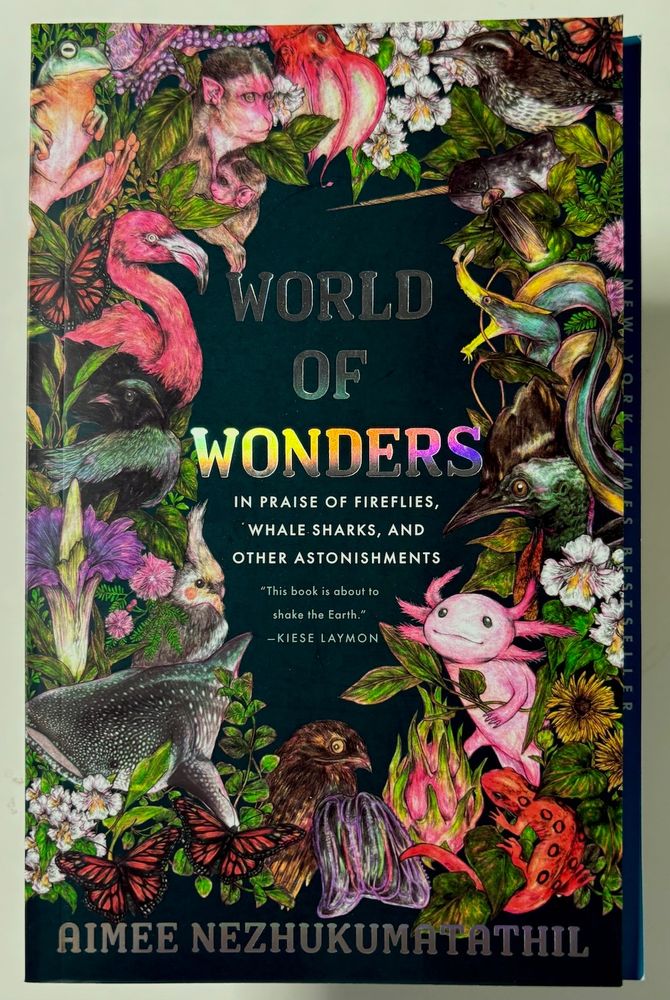 A softcover book colorfully decorated with plants and animals described inside the book - including a Dancing Frog, a Flamingo, a Ribbon Eel, an Axolotl, a Cassowary, Monarch Butterfly, Dwarf Kingfisher, Cactus Wren, a Corpse Flower, a Narwhal and more. The subtitle reads 'In praise of Fireflies, Whale Sharks and Other Astonishments'. All seen against a black background, framing the titles in the center. The main titles iridescent, and change color when the book is turned.