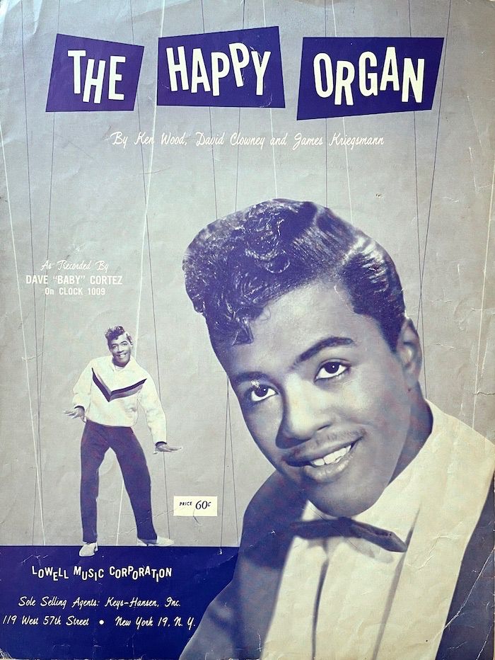 Cover of sheet music 'The Happy Organ, printed in blue; with a photo portrait of doo-wop artist Dave "Baby" Cortez in the foreground. He sports a typical pompadour haircut of the era and wears a thin bow tie. To the left, in the background he can be seen again in slightly a more casual outfit. He appears to be dancing to the beat. 