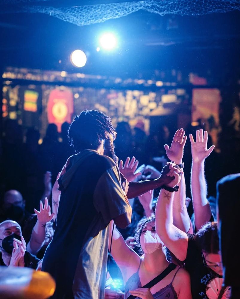 Jer Hunter on stage, mostly in silhouette with their back to the camera. I'm in the lower left, looking up at them with my hands clasped in joy in front of me. Jer is clasping hands with a person in the audience who is looking down, looking pretty emotional. All of the audience in the front by Jer have one arm in the air, reaching out.  