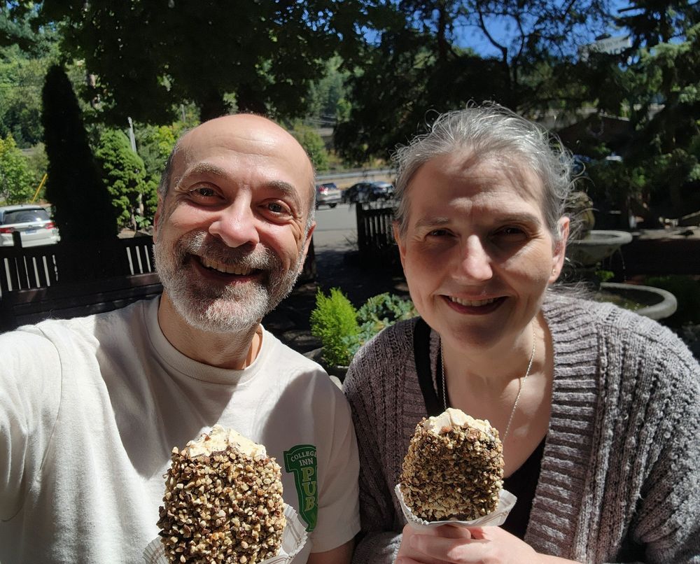 A selfie of my wife and I, both with hand-dipped vanilla ice cream bars covered in dark chocolate and nuts, outside in a sunny garden next to Boehm's Chocolates in Issaquah, WA. A few bites out of each of ours.