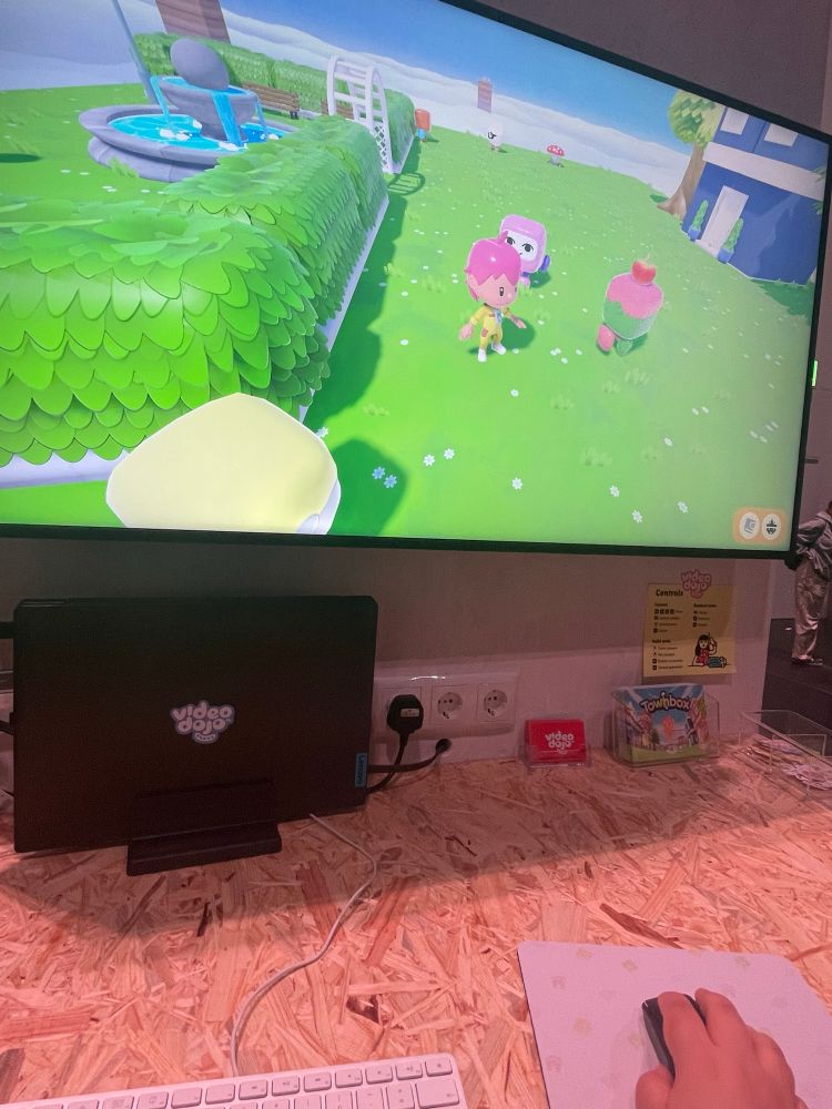 A big PC screen displaying Townbox, an upcoming, in development single-player sandbox town-building game that emphasizes creativity and self-expression. Players take on the role of a trainee town planner, starting with a tiny island and expanding it into a bustling, unique town designed entirely to their vision. On the screen you see the base character; they’re tiny and have pink hair and wear a yellow shirt with matching shorts. The background is a green meadow with some buildings and trees built by the creators in creator mode. The NPCs are all cute little creatures shaped like game boys not unlike gacha capsule toys. It’s a very cute and vibrant game.