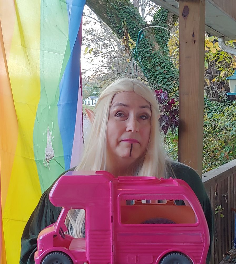 A white woman wearing a long blonde wig and elf ears holds a bright pink toy bus
