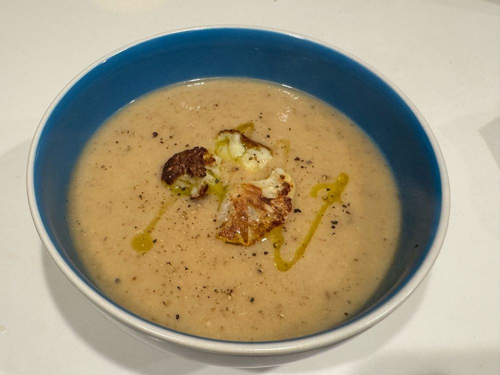 a blended creamy and cream-colored soup in a blue bowl and garnished with olive oil, some roasted cauliflower and crushed pepper