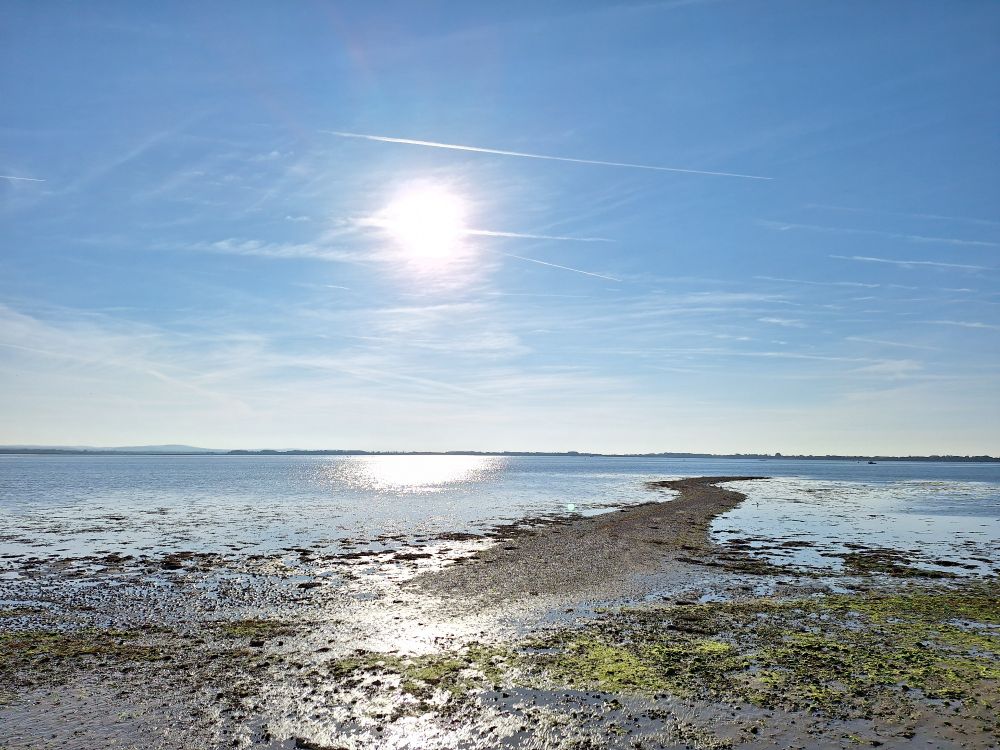 Image shows Langstone harbour at near high tide under a sunny sky. A shingle spit leads out to sea in the centre right of the frame 
