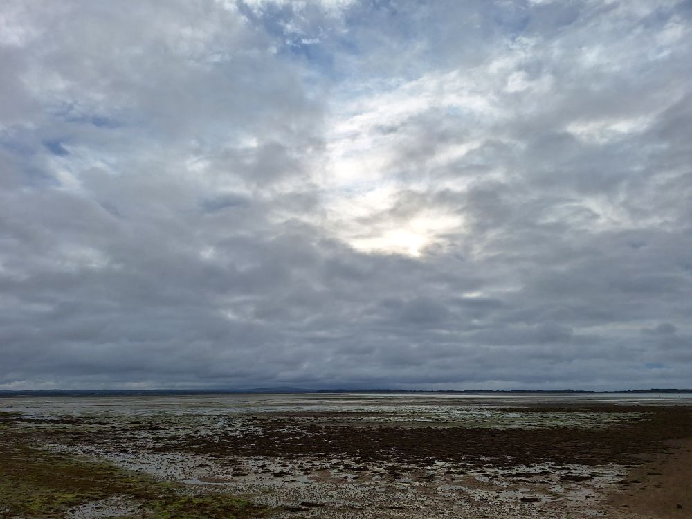 Image shows Langstone harbour at low tide under a mainly cloudy sky. The sun is shining through the clouds 