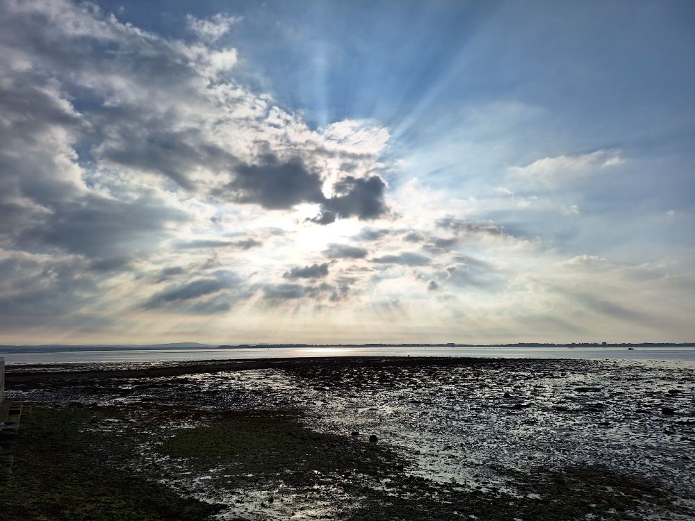 Image shows Langstone harbour at mid tide under a partially cloudy sky. Sun rays shine out from behind the clouds 
