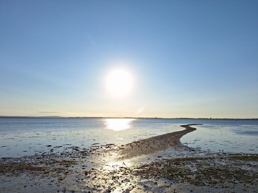 Image shows Langstone harbour at near high tide under a, sunny sky. A shingle spit leads out to sea in centre-right of the frame 
