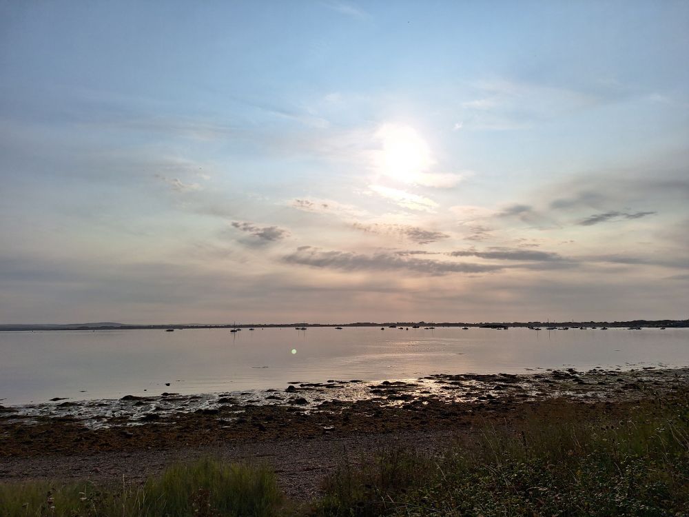 Image shows Langstone harbour at near high tide under a mainly clear sky. A few clouds line the horizon 