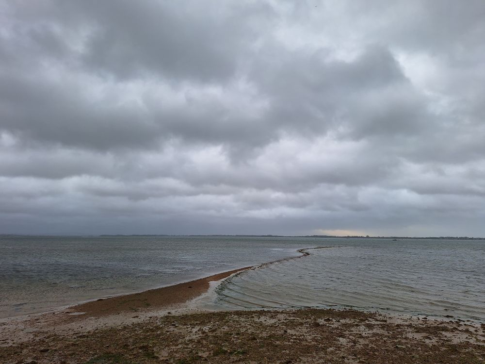 Image shows Langstone harbour at near high tide under a stormy sky. A shingle spit leads out to sea from the left-hand side of the frame 