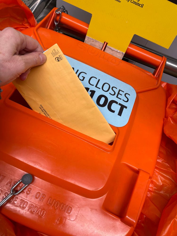 An orange envelope containing local authority voting papers is being dropped into the ‘postal slot’ of an official orange ballot wheelie bin. 