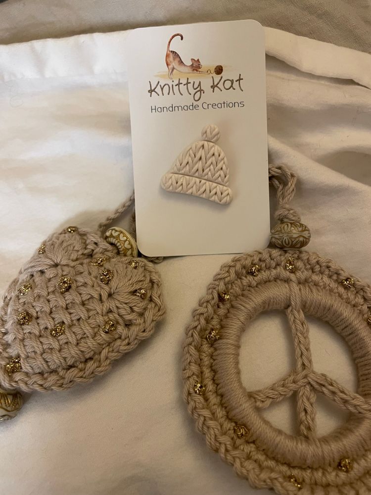 Two handmade Christmas decorations - heart and a peace sign - in beige yarn with beads and golden highlights. And a ceramic pin in the form of a knitted beanie, complete with tiny pompom. 