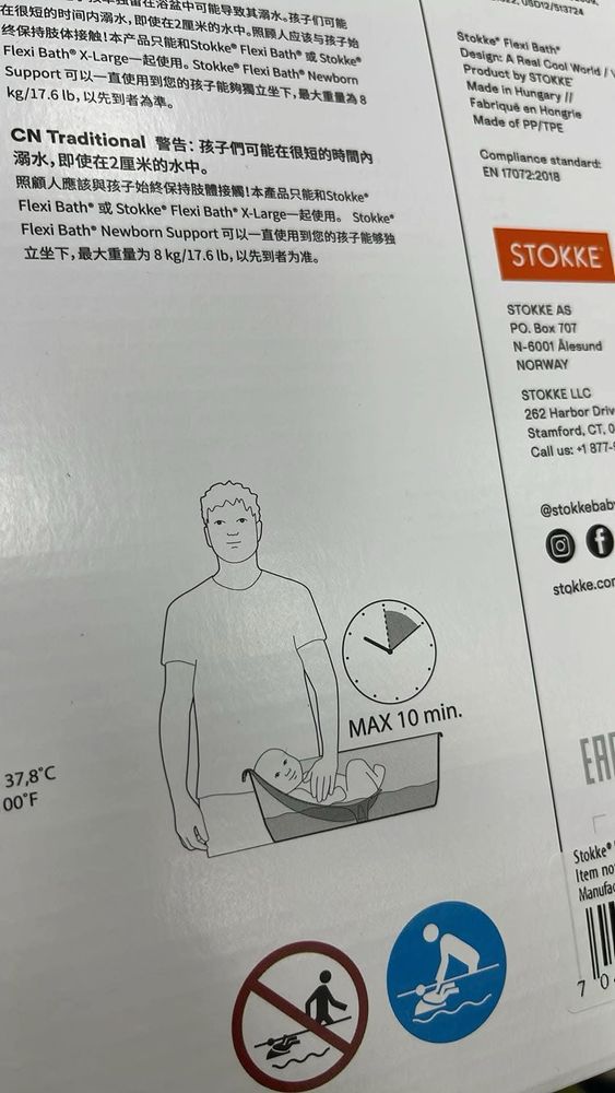 Short description:
Packaging for a Stokke Flexi Bath, featuring safety warnings in multiple languages, a diagram of an adult bathing a baby, a “Max 10 min.” time limit, and temperature recommendations.

Detailed description:
The image shows the packaging of a Stokke Flexi Bath, a collapsible baby bathtub. The packaging includes safety warnings in multiple languages, including Chinese and English, advising caregivers to closely monitor babies during bath time to prevent drowning risks. A black-and-white illustration depicts an adult standing beside a baby lying in the Flexi Bath with a supportive insert. A clock icon with “Max 10 min.” indicates a recommended bath duration limit. The suggested water temperature of 37.8°C (100°F) is also displayed. Below, two symbols provide additional safety guidance: one red-circle prohibition sign with a figure walking into deep water, indicating not to leave the baby unattended or use the bath unsafely, and another blue-circle symbol of an adult holding a baby in shallow water, emphasizing proper supervision. The packaging also includes branding elements for Stokke, company contact details, and manufacturing information.
