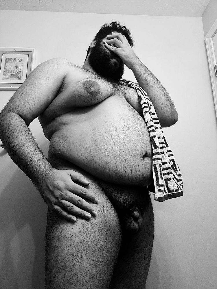 Chubby man covers his face with a hand while the other rests on his thigh. His nude body is there for your viewing pleasure.