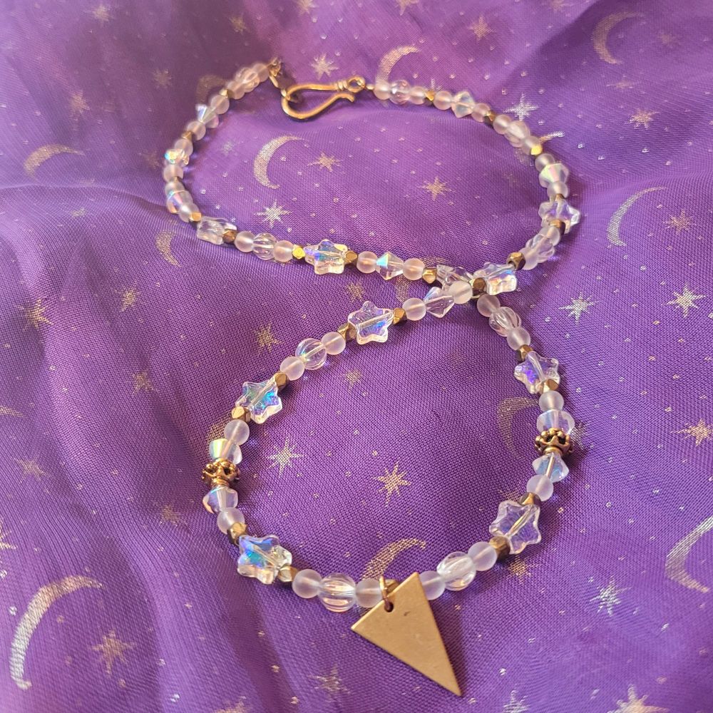 A white and gold beaded necklace with clear round beads, faceted gold beads and shimmery clear star beads looped around itself with a golden hook clasp. A brass triangle pendant hangs at the bottom.