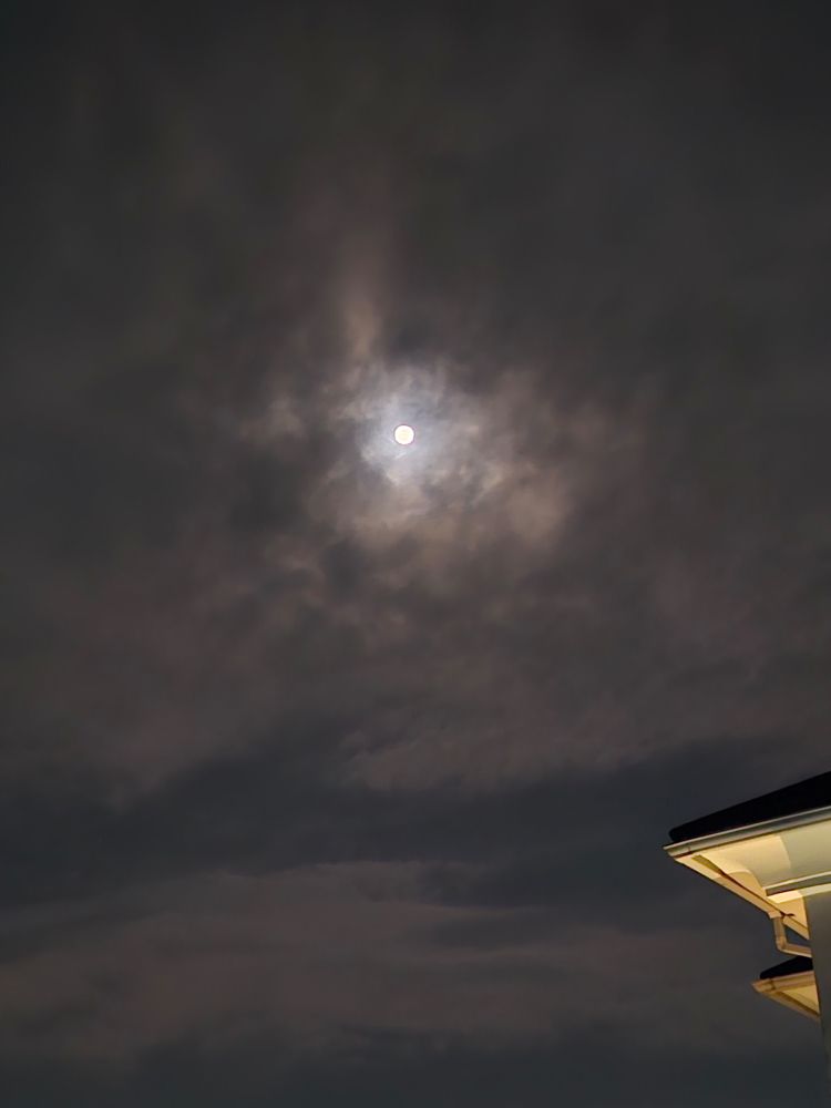 A full moon light a jewel well lit in the middle of a grey cloudscape.
Building roof edge visible on the right
