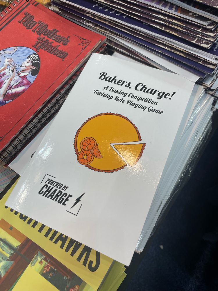 A stack of Bakers, Charge! booklets, a roleplaying game by TTRPGkids. The cover is white with black text and has a pie with a slice being removed.