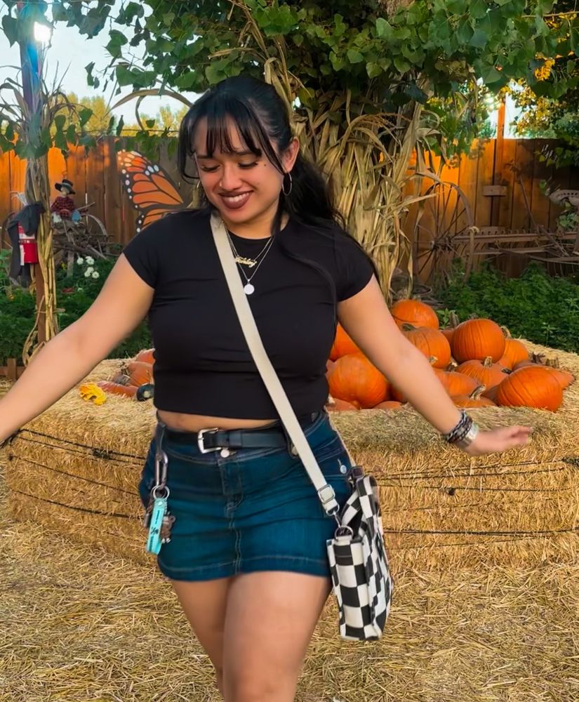 girl is walking in front of pumpkins; its a pumpkin patch. her hair is pulled back into a half updo. she wears a black top, denim skirt, with a checkered bag and guys on her sides. 
