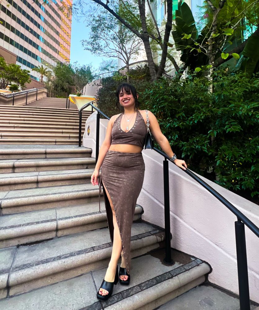 girl posing down a set of stairs, holding onto a rail. shes in LA. her hair is styled into an updo, a brown skirt set, the top a halter, skirt with a slit. black mules. 