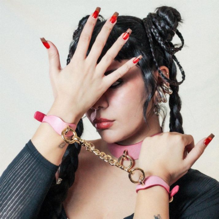 Woman with braided hair and red nails covers face with hand, wearing a pink choker and gold chain-linked wrist cuffs, expressing bold fashion.