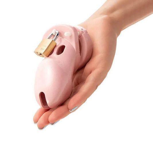A hand holds a pink plastic chastity device with a small brass padlock at the top, designed for male genitalia.