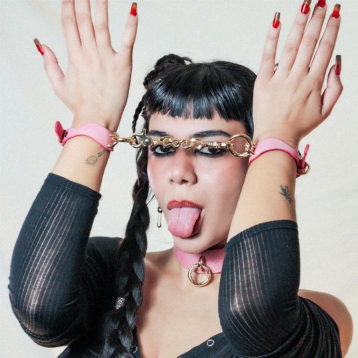A woman with long dark braids and bangs sticks out her tongue. She raises her hands, showcasing pink leather cuffs linked by a chain.