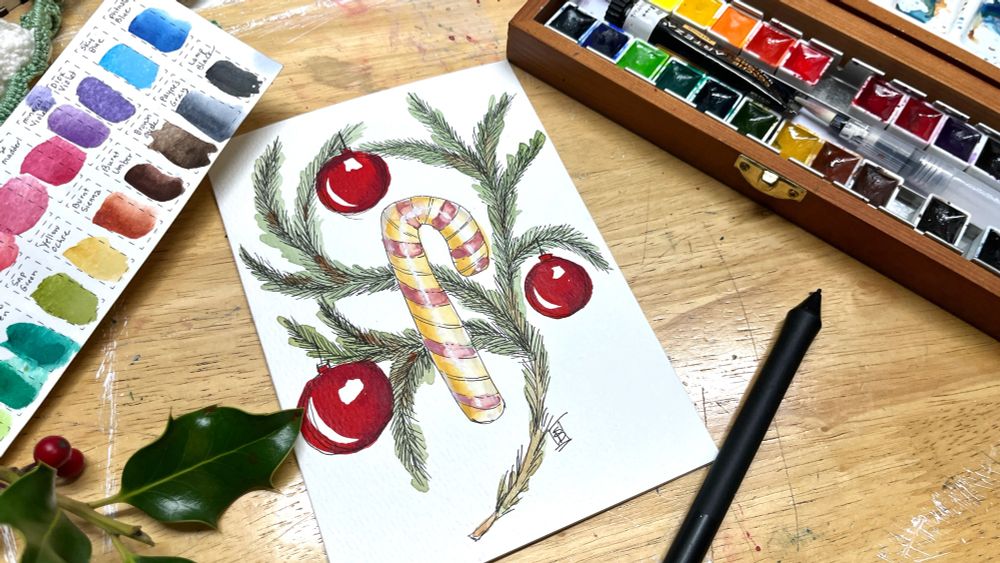 Watercolor palette and colorful swatch card with an ink and watercolor holiday card on a wood background. The holiday card is white with a large pink and cream candy cane on a background of pine boughs and shiny red ball ornaments- painted on a video by Stephanie Bergeron on Deliberately Creative YouTube channel 