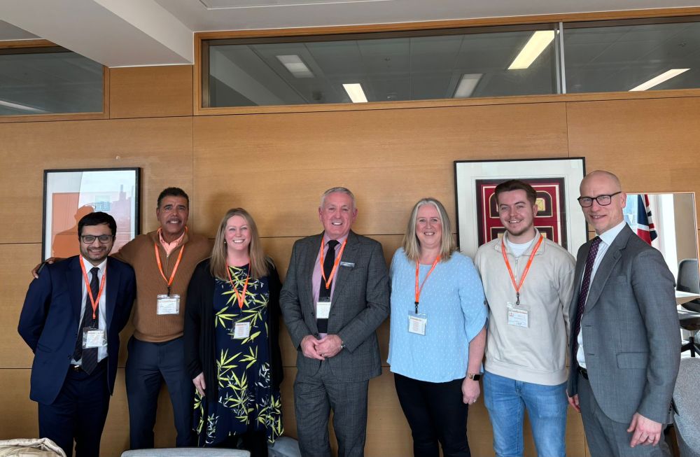 A photo of (left to right) Saqib Bhatti, MP for Meriden, Chris Kamara, Gillian Rudd, Steve Jamieson, Louisa Akers, Mikey Akers and Minister Stephen Kinnock. Everyone is stood up and smiling. The background is a wall of wooden panelling with two framed pictures on the wall and windows at the top.