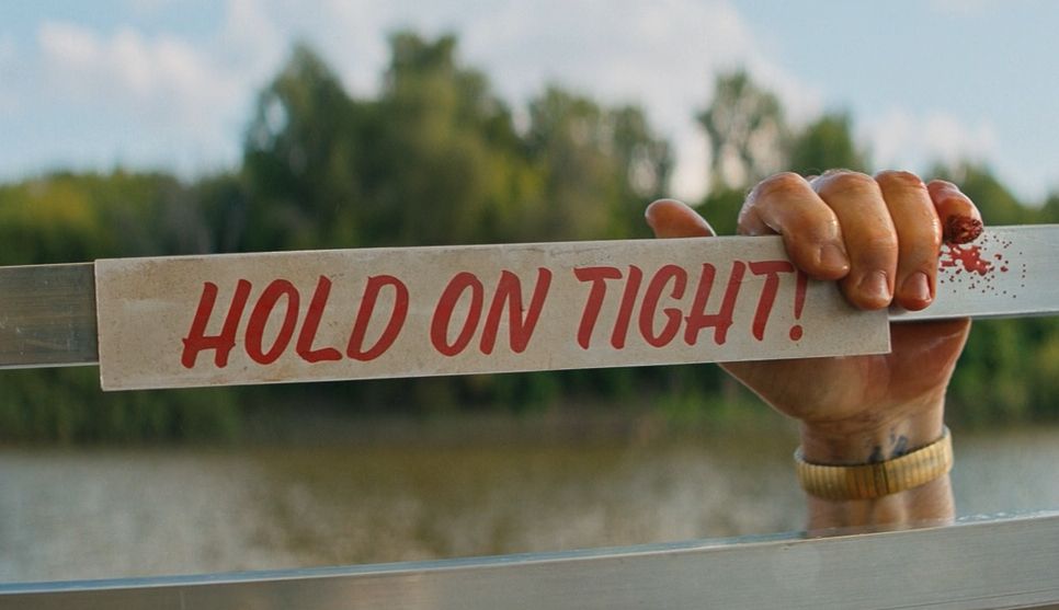 A partial screenshot of the movie Nobody 2 (2025). It shows a metal railing with a white sign stating in red letters, "HOLD ON TIGHT!"  And there's a hand grabbing the railing, as if someone is trying to climb using this railing. But the hand has a severed pinky.