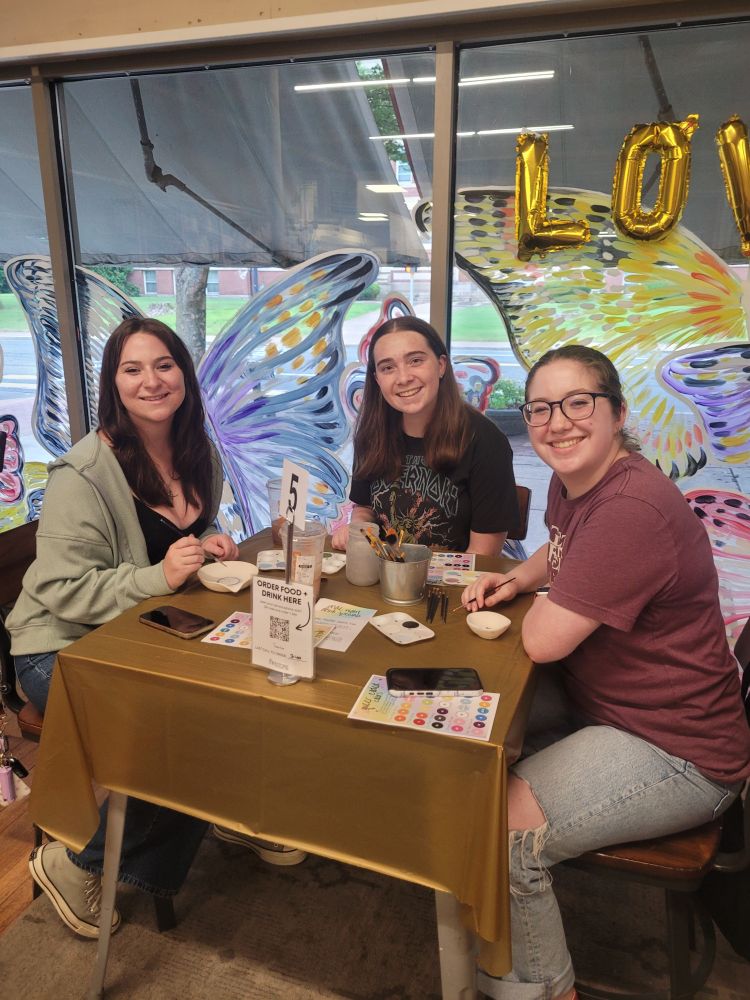 Three friends sit at a table painting ceramics and smiling.