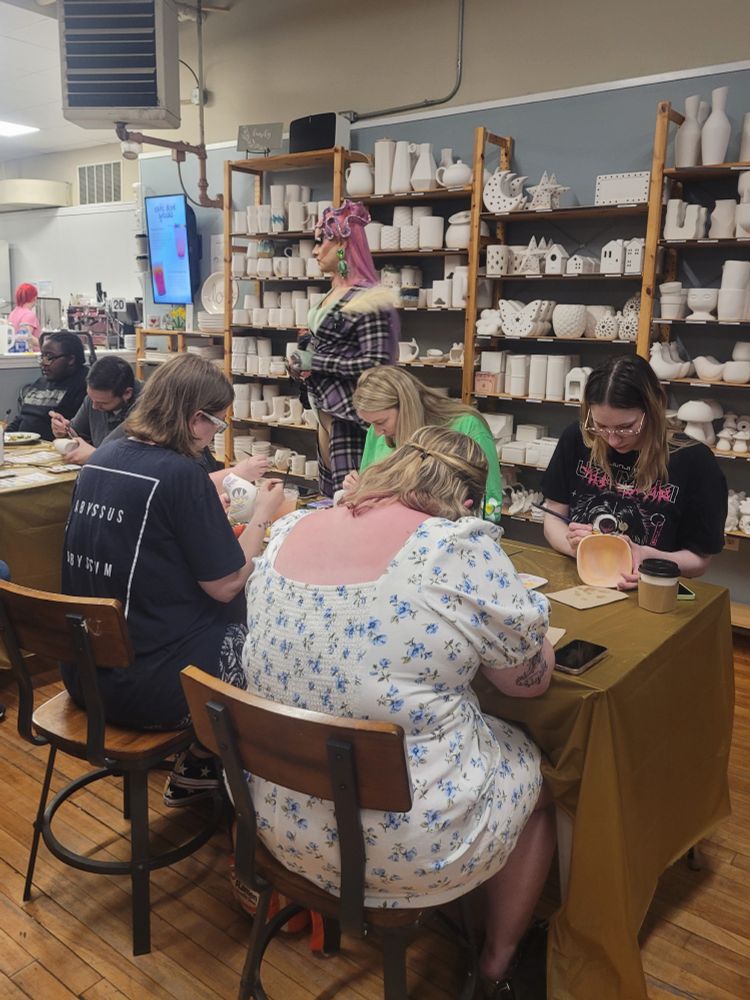 This is a candid shot of four people painting ceramics at a table, while a drag performer stands in the background.