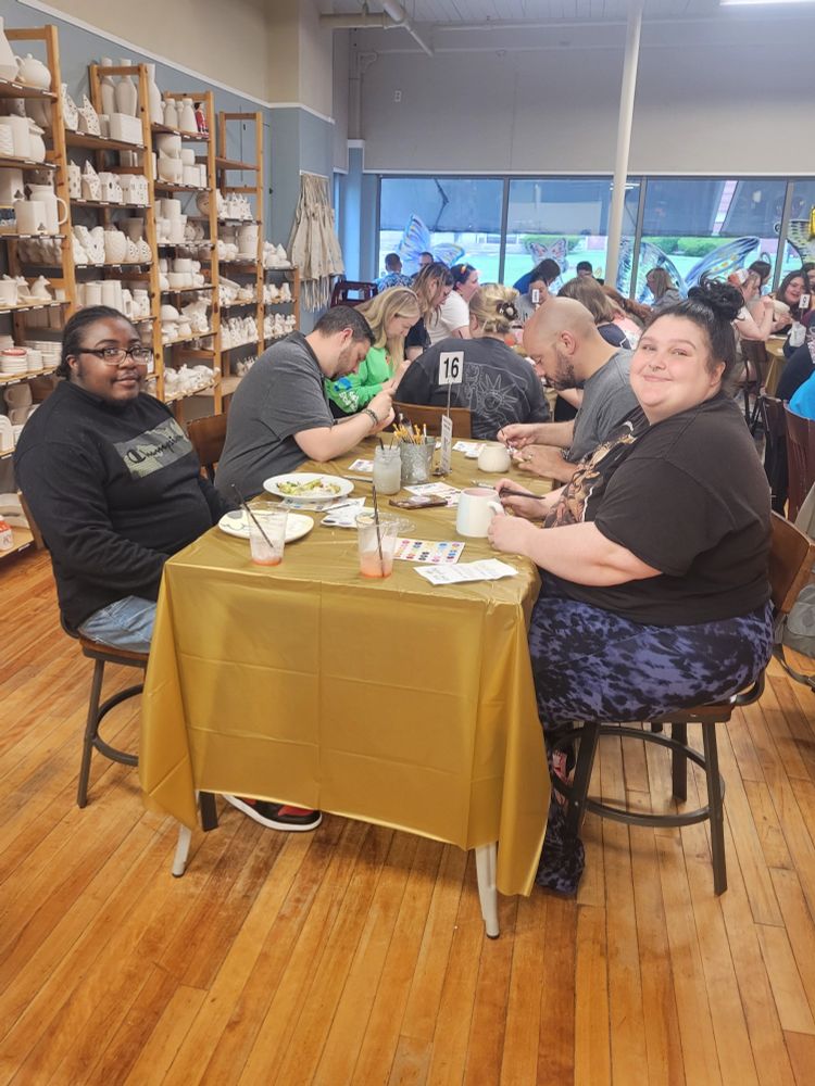 Two friends paint ceramics at a table and smile at the camera.