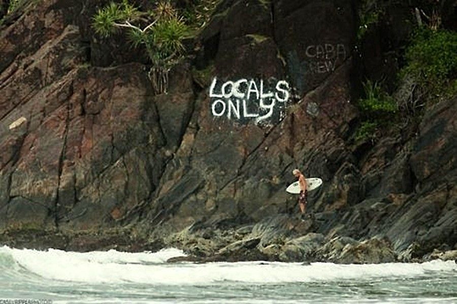 A surfer holding a surfboard on the shore with "Locals Only' graffiti sprayed on the cliff above.