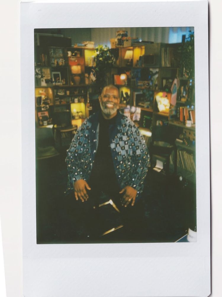 foto em estilo polaroid do cantor Perícles. ele está sentado, sorrindo, usando casaco azul e roupa preta. o cantor está no cenário do tiny desk brasil.