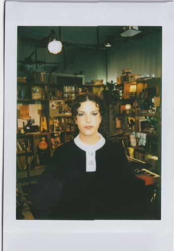 foto em estilo polaroid da cantora Céu. ela está em pé, de cabelo preso, usando roupa preta com detalhes gola e detalhe em branco (parece a toga de um juiz). a cantora está no cenário do tiny desk brasil.