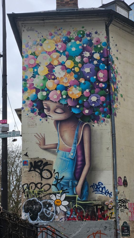 Cette œuvre représente un personnage féminin aux yeux fermés, avec une chevelure afro composée de bulles colorées éclatées, évoquant des fleurs ou des explosions de peinture.  Le personnage porte une salopette bleue et semble apaisé, la main levée dans un geste doux.  Le bas du mur est recouvert de graffitis, de collages et de tags variés.