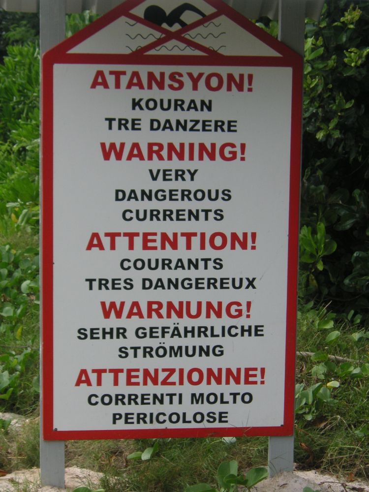 Cette photo montre un panneau d’avertissement multilingue installé sur une zone côtière. En haut, un pictogramme représente une personne nageant dans des vagues, signalant un danger lié à l’eau. 

Le message principal met en garde contre des courants marins très dangereux, et il est traduit en cinq langues : créole seychellois, anglais, français, allemand et italien.