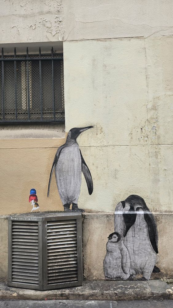 C'est une œuvre de street art représentant trois manchots, dont un grand debout sur une boîte de ventilation et un groupe de deux (un adulte et un plus petit) à droite, peints sur un mur. 