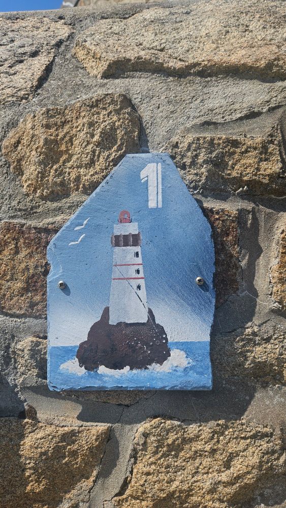 Sur la photo, on voit une plaque de maison en ardoise fixée sur un mur en pierre. La plaque a une forme de maisonnette stylisée, peinte en bleu ciel. Au centre de la plaque, un phare est dessiné posé sur un rocher entouré de vagues. Deux mouettes blanches volent au-dessus de l'eau. Le chiffre 1 est inscrit en haut de la plaque.