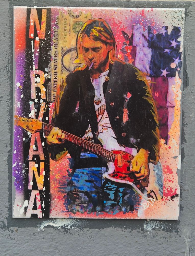 Ce street art représente Kurt Cobain. 

L’artiste utilise une combinaison de couleurs éclatantes – rose, violet, orange et bleu.

La silhouette centrale montre le  guitariste en pleine attitude rock, cigarette aux lèvres, vêtu d’un gilet sombre et d’un t-shirt clair.

À gauche, le mot NIRVANA apparaît en grandes lettres verticales.

En arrière-plan, on distingue des éléments graphiques tels qu’un billet de banque et diverses couches colorées qui donnent de la profondeur à l’ensemble.
