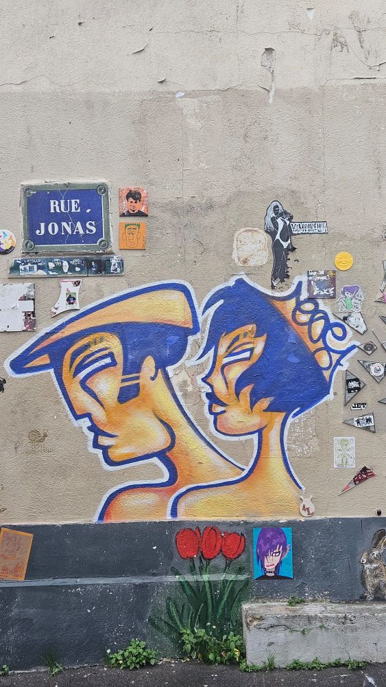 Le street art principal de cette photo représente deux visages de profil, côte à côte, peints sur un mur beige. 

Leurs visages sont  allongés, peints dans des tons chauds de jaune et d’orange, avec des ombres marquées.

Le style est à la fois graphique et stylisé, avec des lignes fluides et des contours épais de couleur bleu foncé. 

