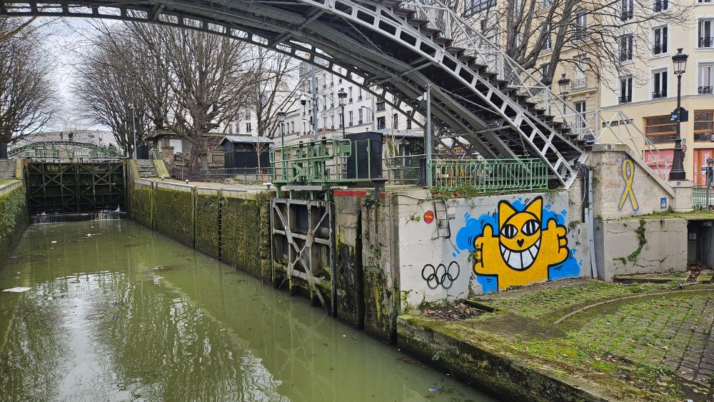 Cette photo montre une écluse du Canal Saint-Martin dans le 10ᵉ arrondissement, sous une passerelle métallique typique du quartier. 

Sur le mur, un chat jaune orangé qui se trouve de face arbore un énorme sourire. 