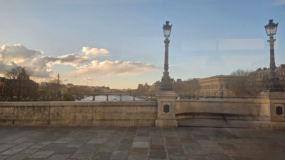La photo prise depuis un pont offre une vue étendue sur la Seine. 

Le ciel est parsemé de nuages cotonneux qui accrochent les rayons du soleil couchant, créant un contraste agréable avec le ciel bleu pâle.
 
Au centre, la Seine s'écoule, traversée par plusieurs ponts élégants. 

Sur la rive opposée, on aperçoit une architecture classique parisienne avec des bâtiments alignés. 

À l'extrême gauche, on distingue clairement la silhouette emblématique de la Tour Eiffel. 

Le premier plan est dominé par une large balustrade en pierre sombre, surmontée de lampadaires à l'ancienne, richement décorés, typiques de l'éclairage public parisien.

Plusieurs personnes sont visibles sur le trottoir dallé à droite.
