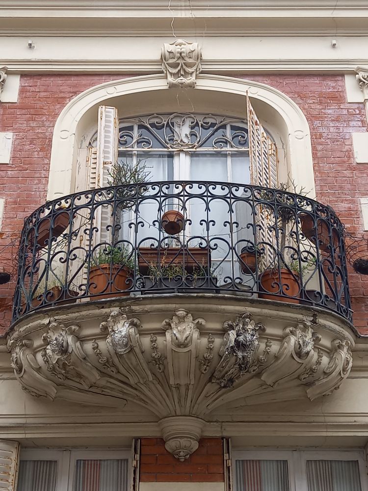 Cette photo montre un balcon orné en fer forgé sur une façade en briques rouges.
Le balcon est soutenu par des consoles sculptées.

Plusieurs pots de plantes sont posés sur le balcon. 
Au-dessus, une fenêtre cintrée avec des volets entrouverts donne sur l'intérieur. 