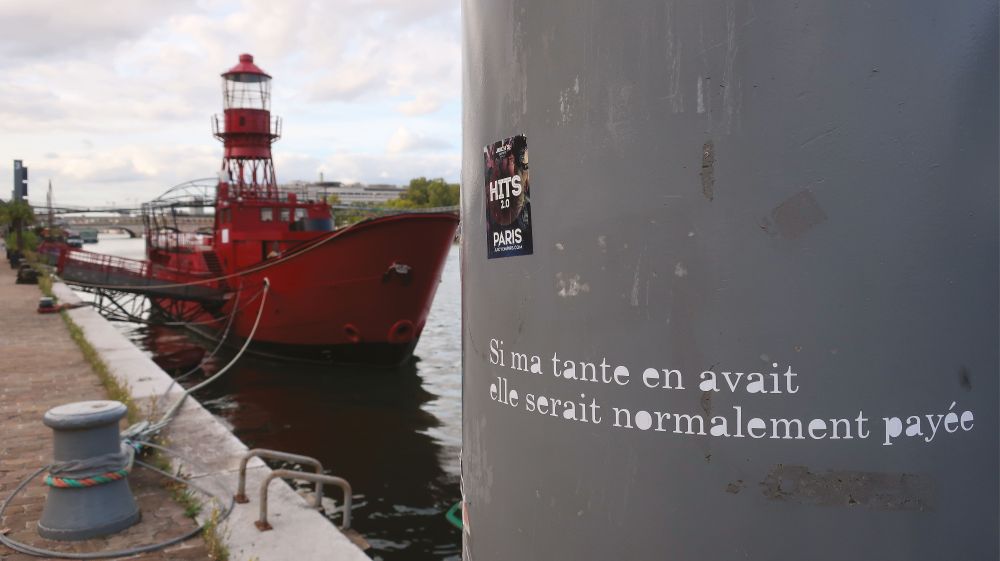 Un bateau-phare rouge vif est amarré le long d'un quai, avec des bittes d'amarrage grises. 
Au premier plan, sur la droite, on voit un poteau métallique gris qui sert de support à un message écrit en grand lettrage blanc :
"Si ma tante en avait
elle serait normalement payée".