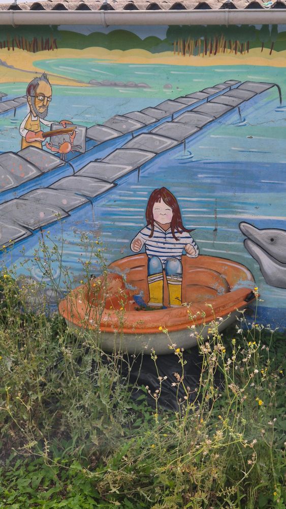 On voit une scène marine peinte sur un mur, avec un décor coloré : l’eau, un ponton, un ostréiculteur barbu en tablier jaune qui cherche à faire fuir un gros crabe qui est sur ses poches et au loin des arbres sur la rive. Au premier plan, la fresque devient très originale parce qu’elle mélange peinture et réel : un petit canot orange, bien réel cette fois, est accolé au mur et prolongé par le dessin. Sur la partie peinte du canot, une fillette en bottes jaunes et marinière est représentée assise, donnant l’impression qu’elle est installée dedans. À côté d’elle, on devine la tête souriante d’un dauphin peint qui sort de l’eau. Elle est incroyable cette fresque : Le contraste entre le canot tangible et la fresque peinte crée une illusion très ludique, comme si la barque sortait vraiment du mur et que la petite fille s’apprêtait à voguer dans cet univers marin plein de vie. 