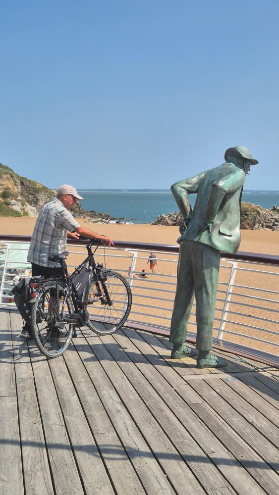 Sur cette photo, on voit un cycliste, vêtu d'une chemise à carreaux et d'une casquette claire, tenant son vélo et regardant dans la même direction que monsieur Hulot.

En effet, à sa droite se trouve la célèbre statue en bronze de Monsieur Hulot, personnage créé par Jacques Tati, reconnaissable à sa posture légèrement penchée en avant, les mains derrière le dos, portant un chapeau et fumant.

La scène se déroule sur une promenade en bois surplombant une plage de sable doré, avec la mer en arrière-plan sous un ciel bleu dégagé.
