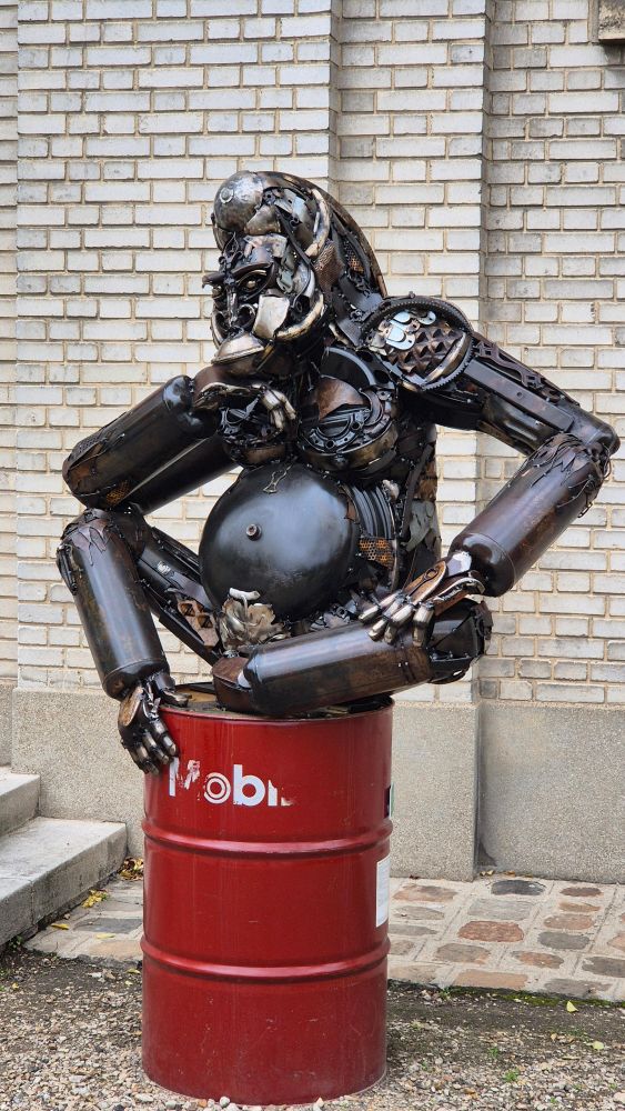 Cette œuvre est une sculpture représentant un gorille entièrement créée à partir de pièces de métal recyclées, ce qui s'inscrit dans le mouvement du "scrap art" (art de la récupération).

Le gorille est représenté dans une posture assise. Son regard semble pensif ou introspectif.

La sculpture est un assemblage soudé de nombreuses pièces mécaniques et métalliques de récupération.

L'ensemble de la sculpture repose sur un fût de pétrole rouge vif de la marque "Mobil", qui lui sert de socle.


