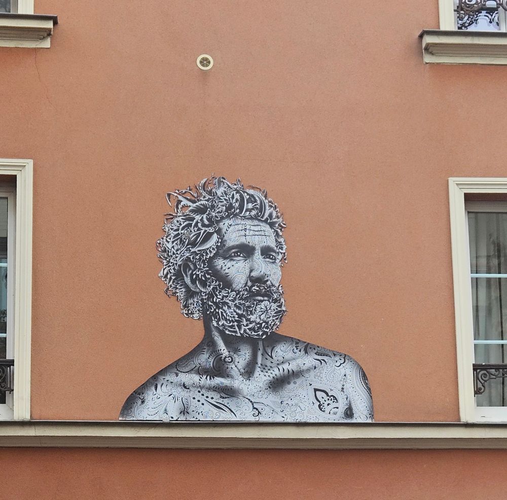 Cette image présente une œuvre de street art collée sur la façade d'un immeuble de couleur terracotta.

L'œuvre représente le buste d'un homme barbu, dans un style qui évoque les figures mythologiques, avec une touche résolument moderne.

Il est de couleur monochrome (gris, blanc et noir), créant un contraste saisissant avec la couleur chaude du mur.

L'homme est couvert de fins tatouages qui recouvrent son buste et pourvu d’une chevelure et d’une barbe fleuries.
