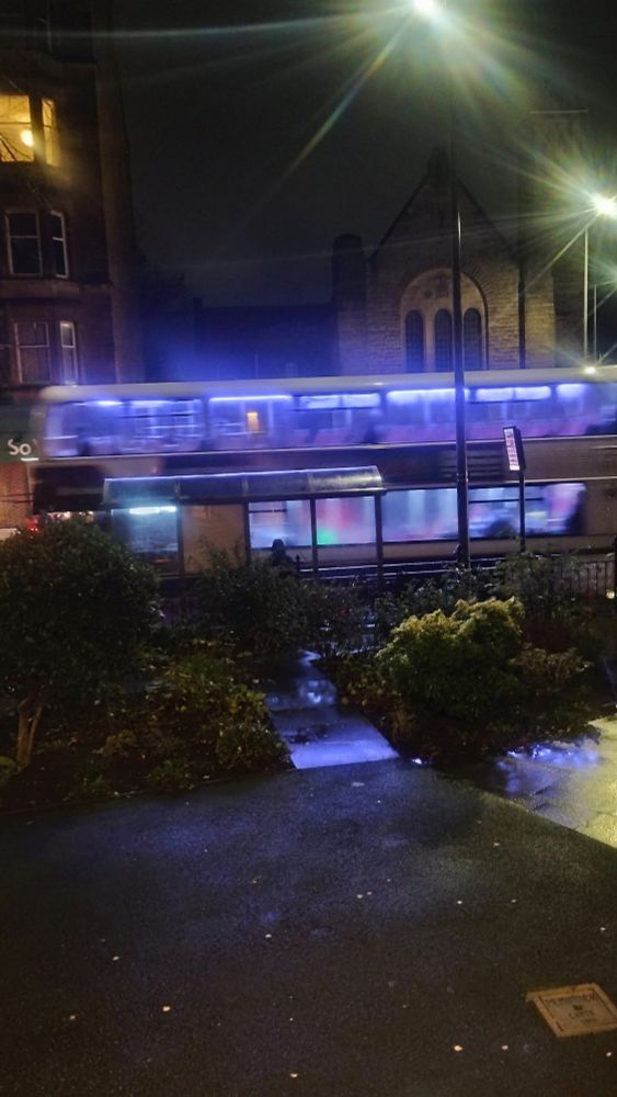 Looking across the garden in front of Christ Church at night, as a double decker bus pulls up to the stop on the pavement beyond 