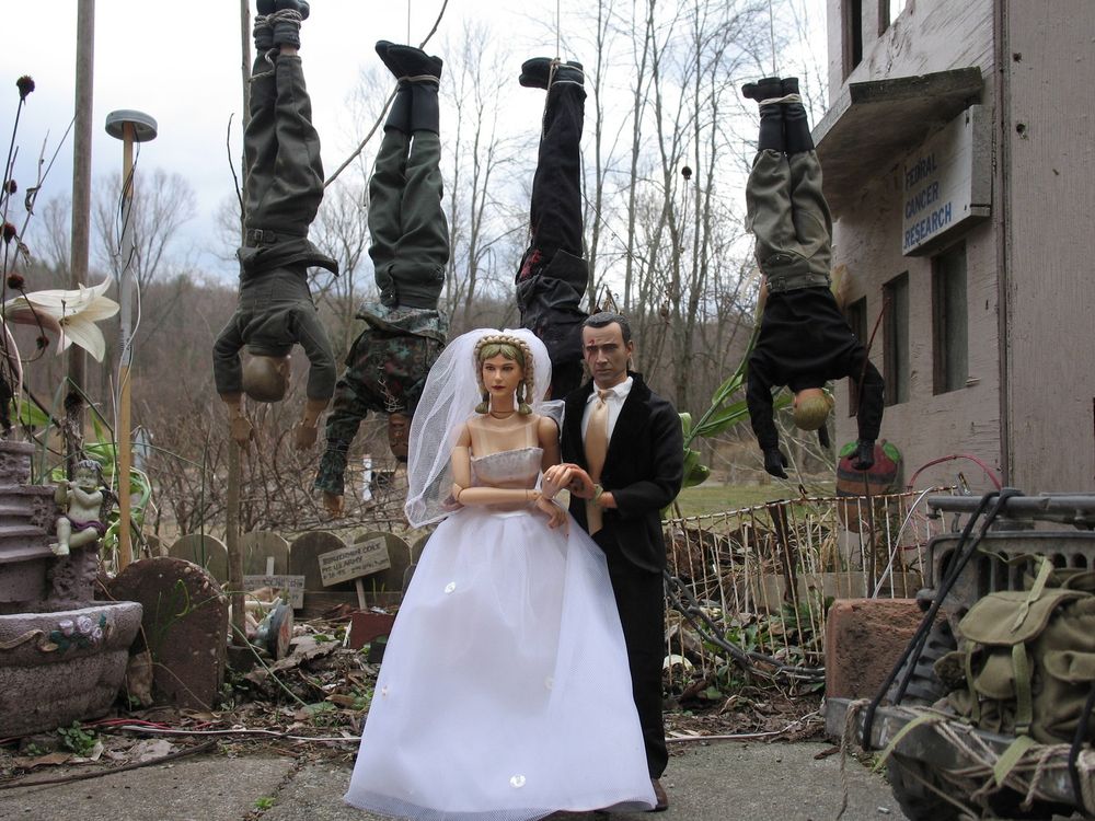 photo of GI Joe dolls hanging by their feet behind a wedding photo of a Barbie doll and a GI Joe by Mark Hogan camp from his book "Welcome to Marwencol"